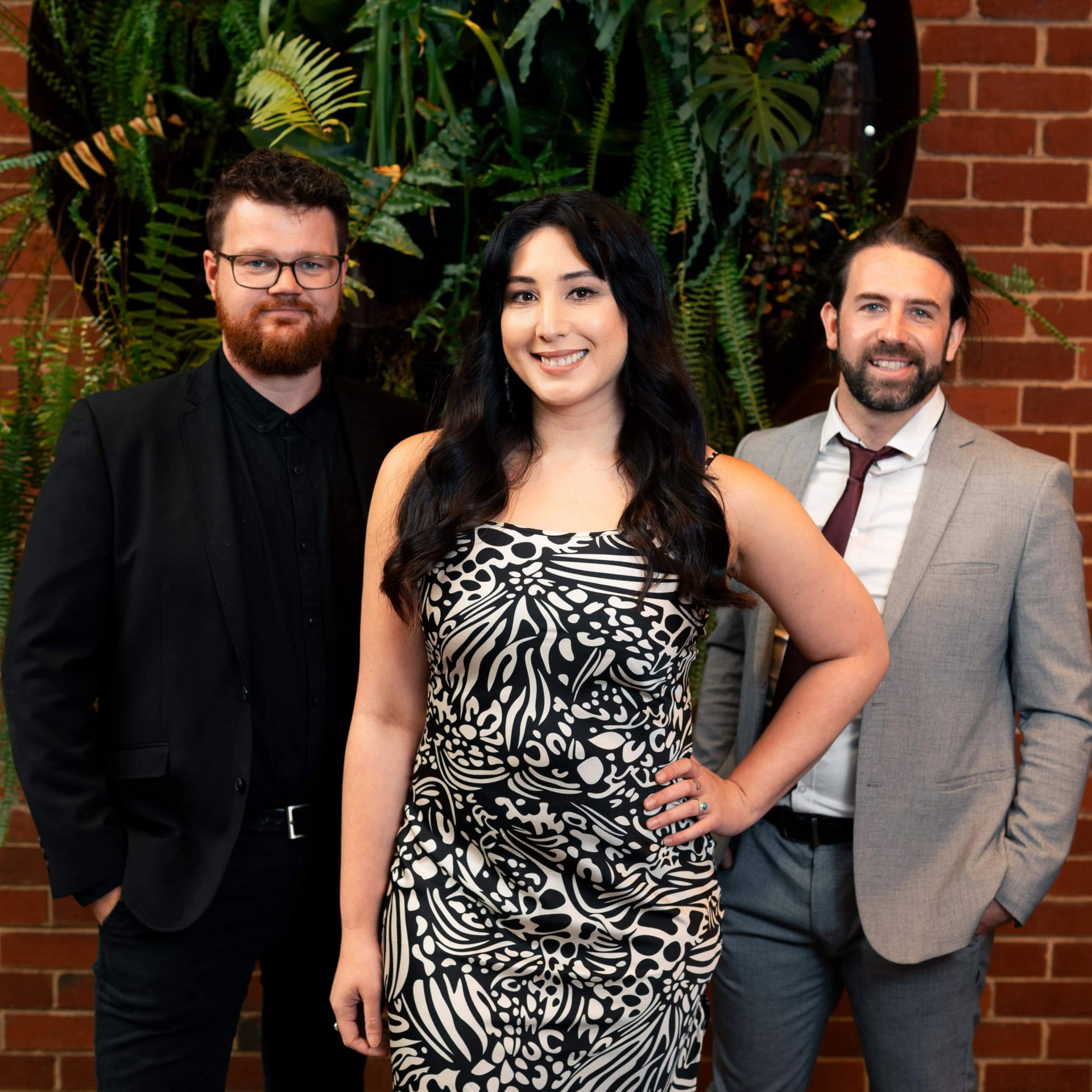 Three people pose in front of a green plant wall. Woman in dress stands centered, flanked by men in suits.