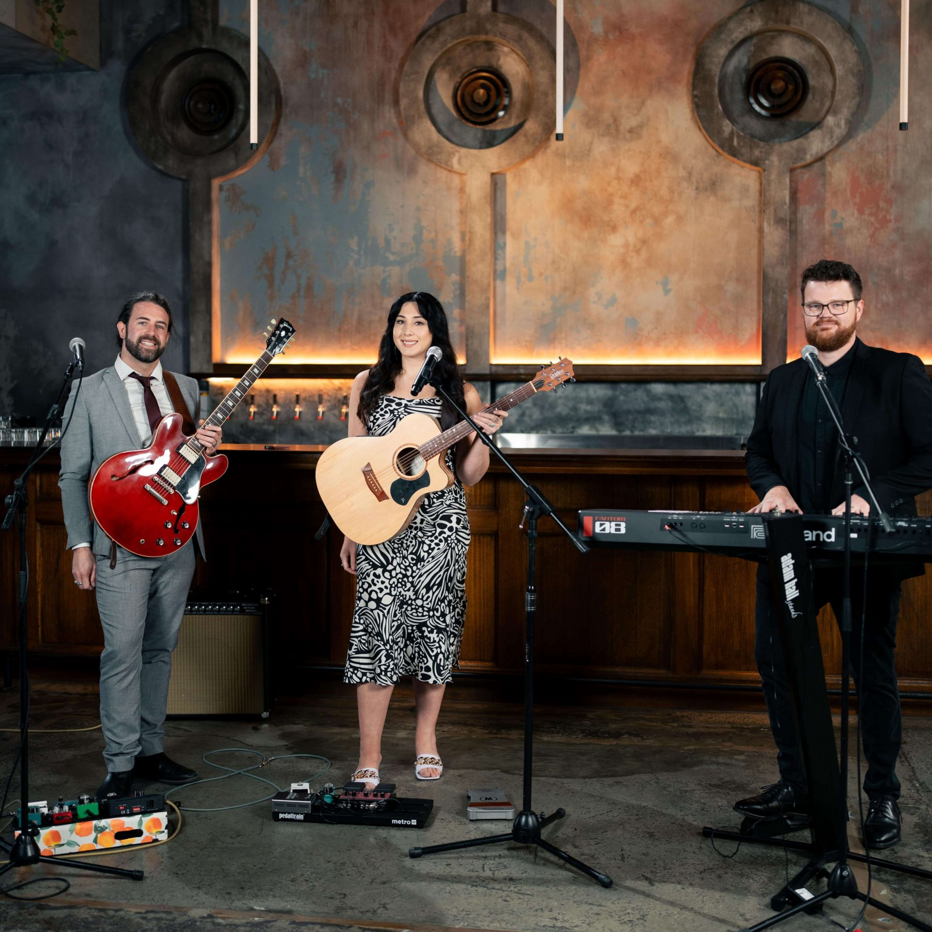 Three musicians performing on a stage: a guitarist, a vocalist with an acoustic guitar, and a keyboardist.