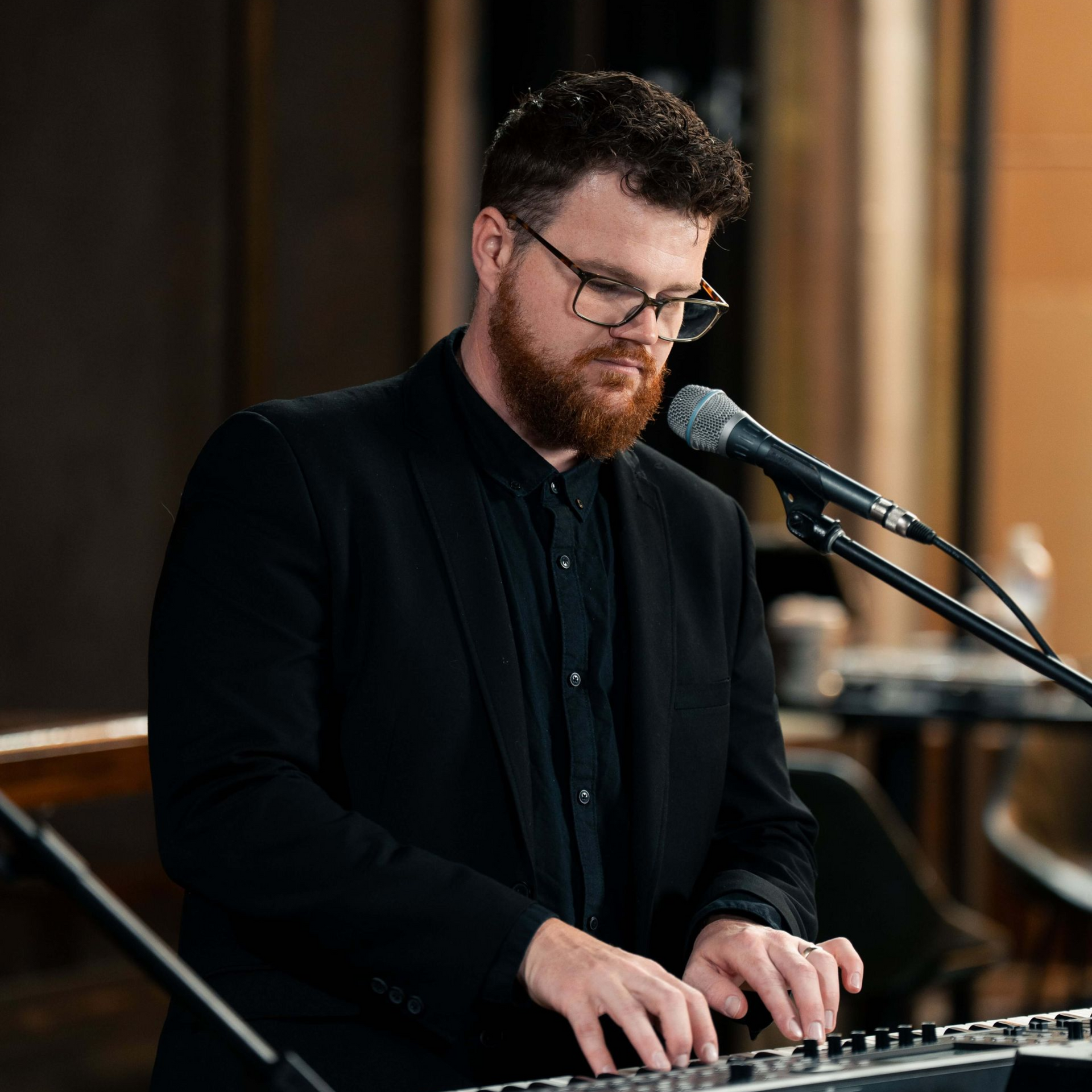 Man with glasses plays a keyboard, singing into a microphone. He wears a black suit in a warmly lit room.
