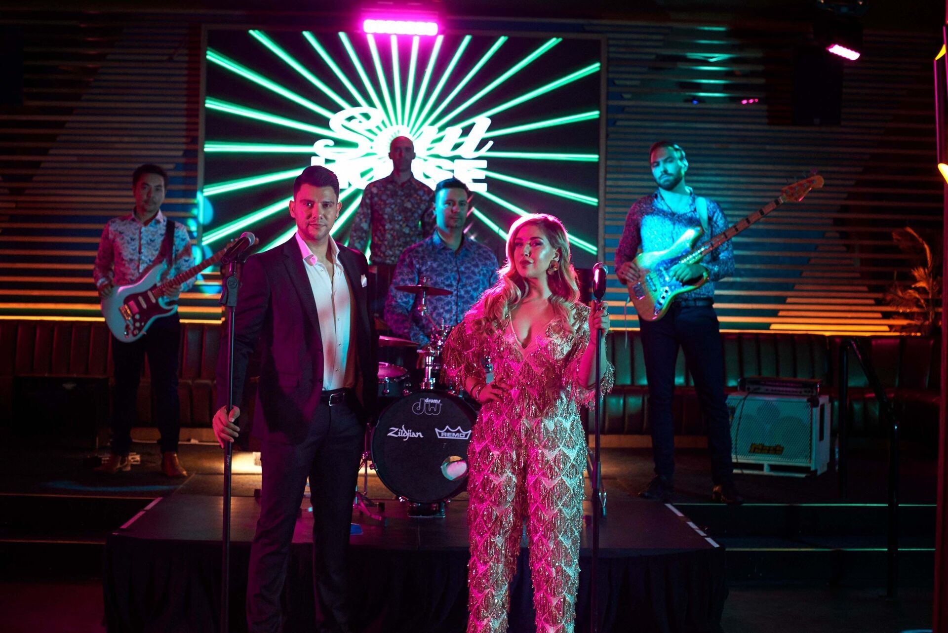 Band performing on stage with neon sign background. Singer in sequined jumpsuit and musician in suit are featured.