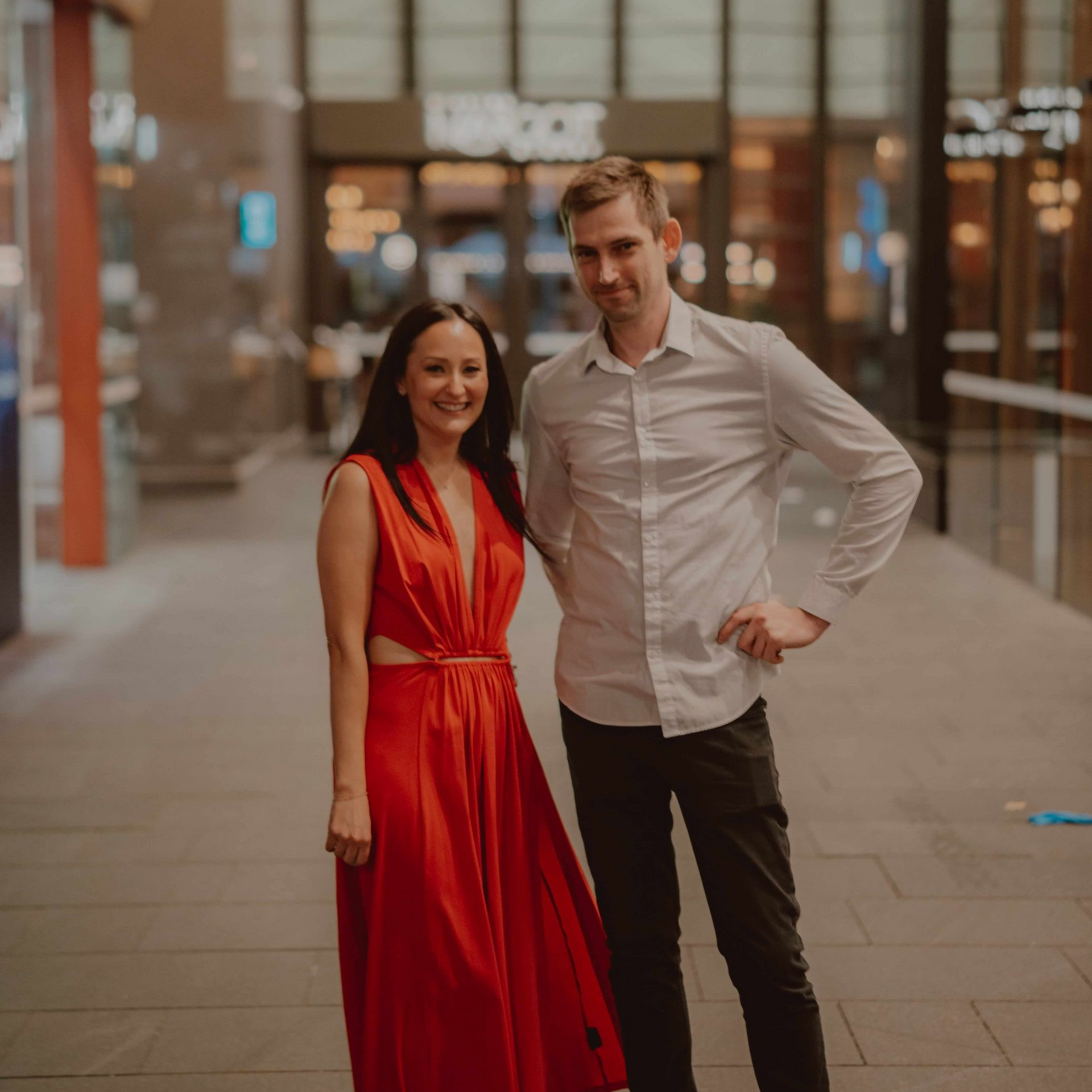 A smiling woman in a red gown and a man in a white shirt and dark pants stand together in a dimly lit, modern hallway.