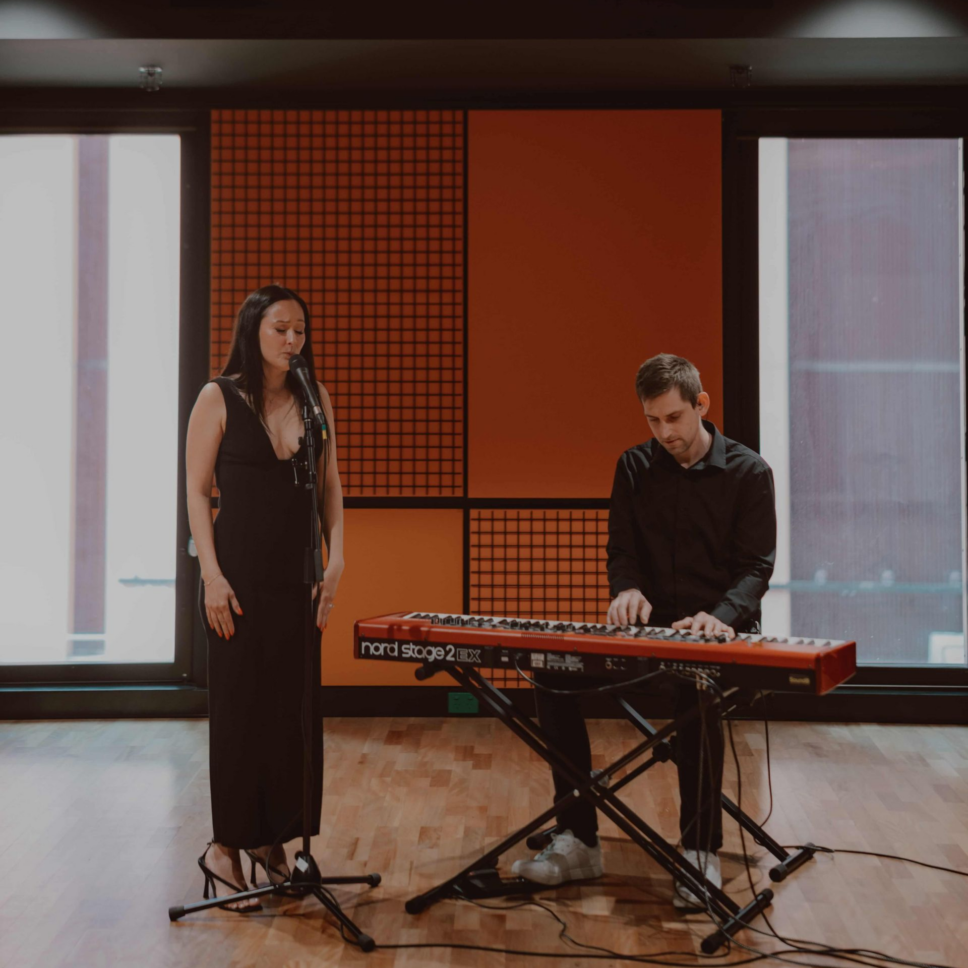A woman sings into a microphone as a person plays a red Nord Stage 2 keyboard in front of an orange geometric wall.