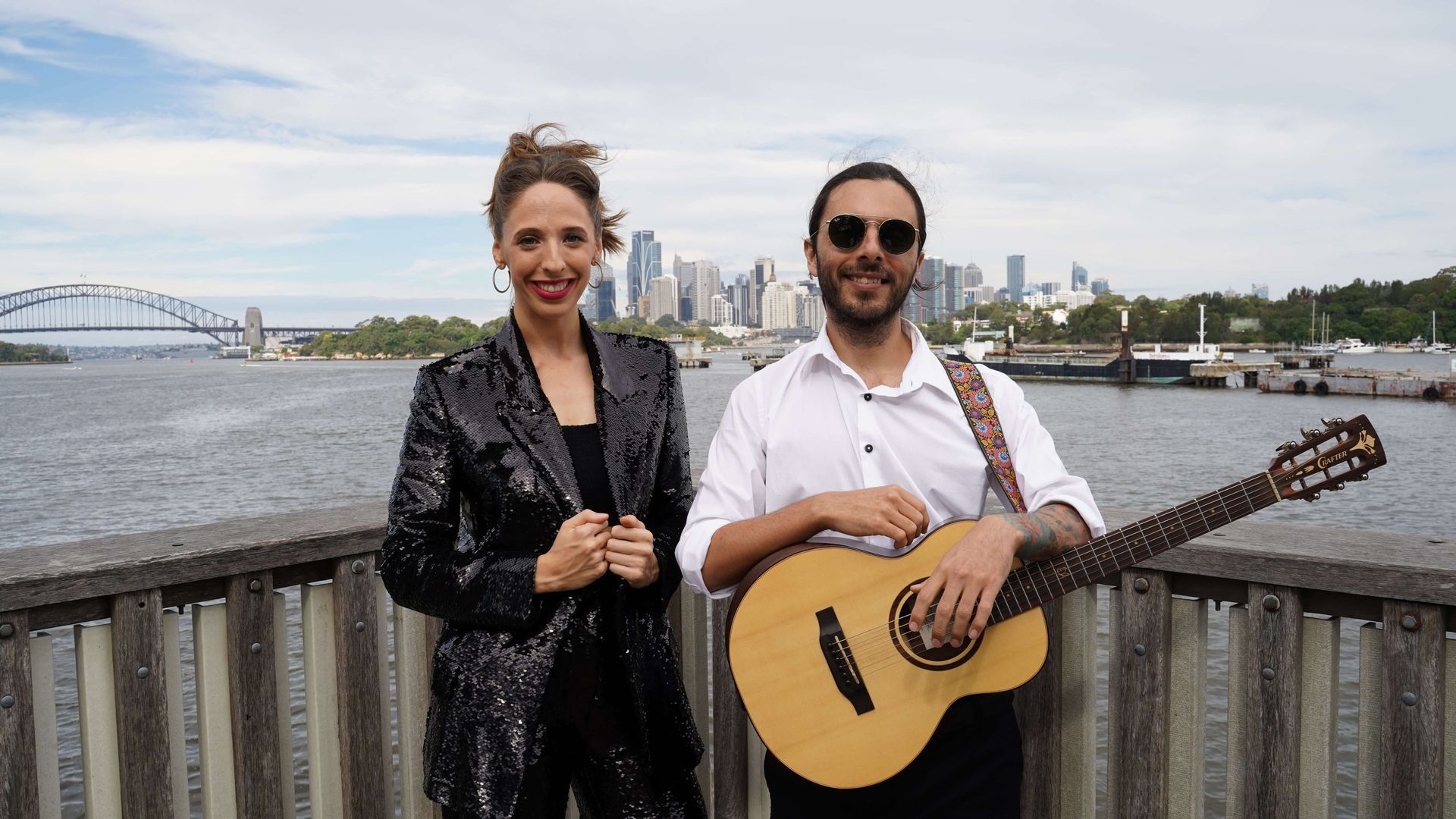 A person in a black sequined jacket and a person playing an acoustic guitar stand smiling before a harbor and city skyline.