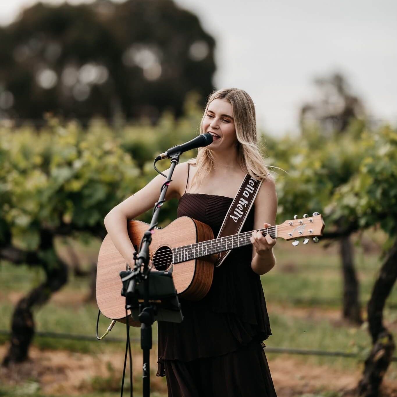 Woman playing guitar and singing at vineyard, wearing a black dress.