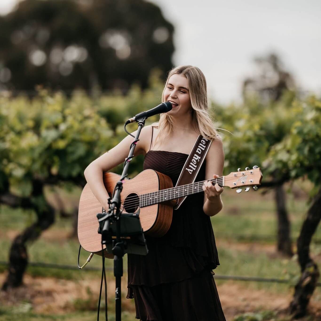Woman playing acoustic guitar and singing into a microphone outdoors in a vineyard.
