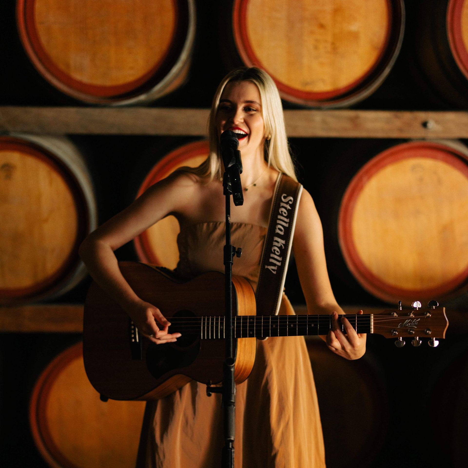 Woman playing acoustic guitar, singing into a microphone in a room with wine barrels.