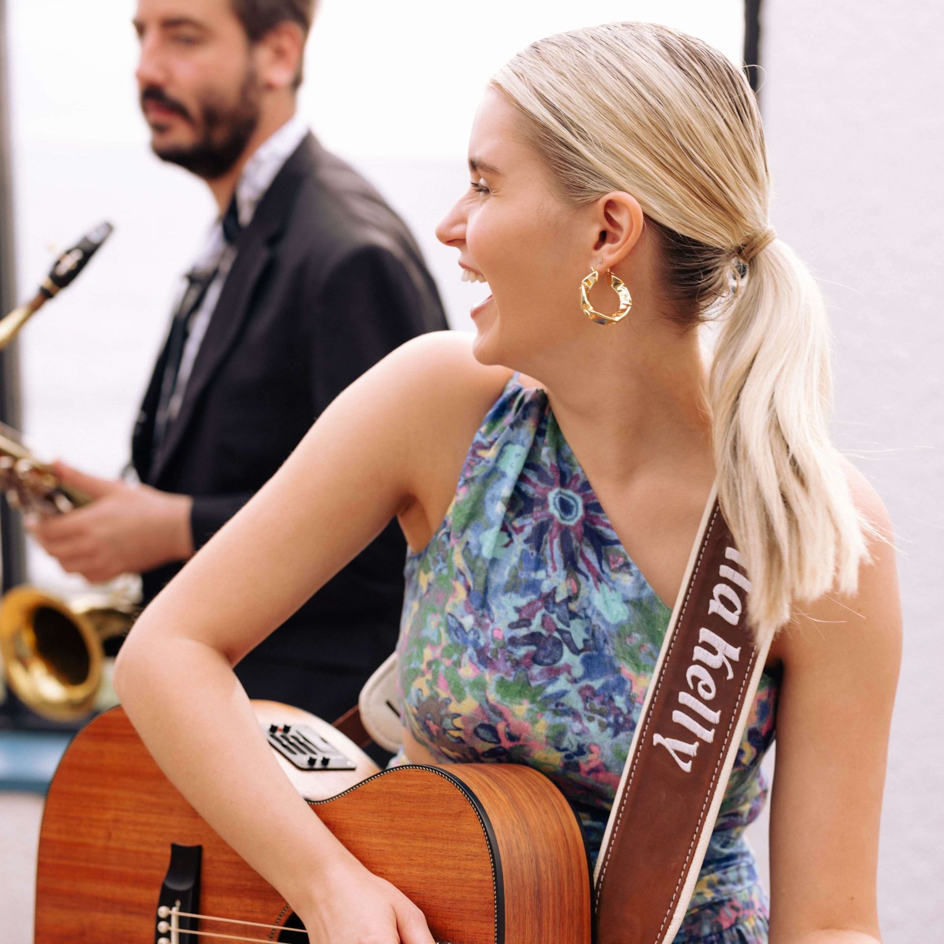 Woman with guitar smiles, performing alongside a saxophonist in a sunny outdoor setting.