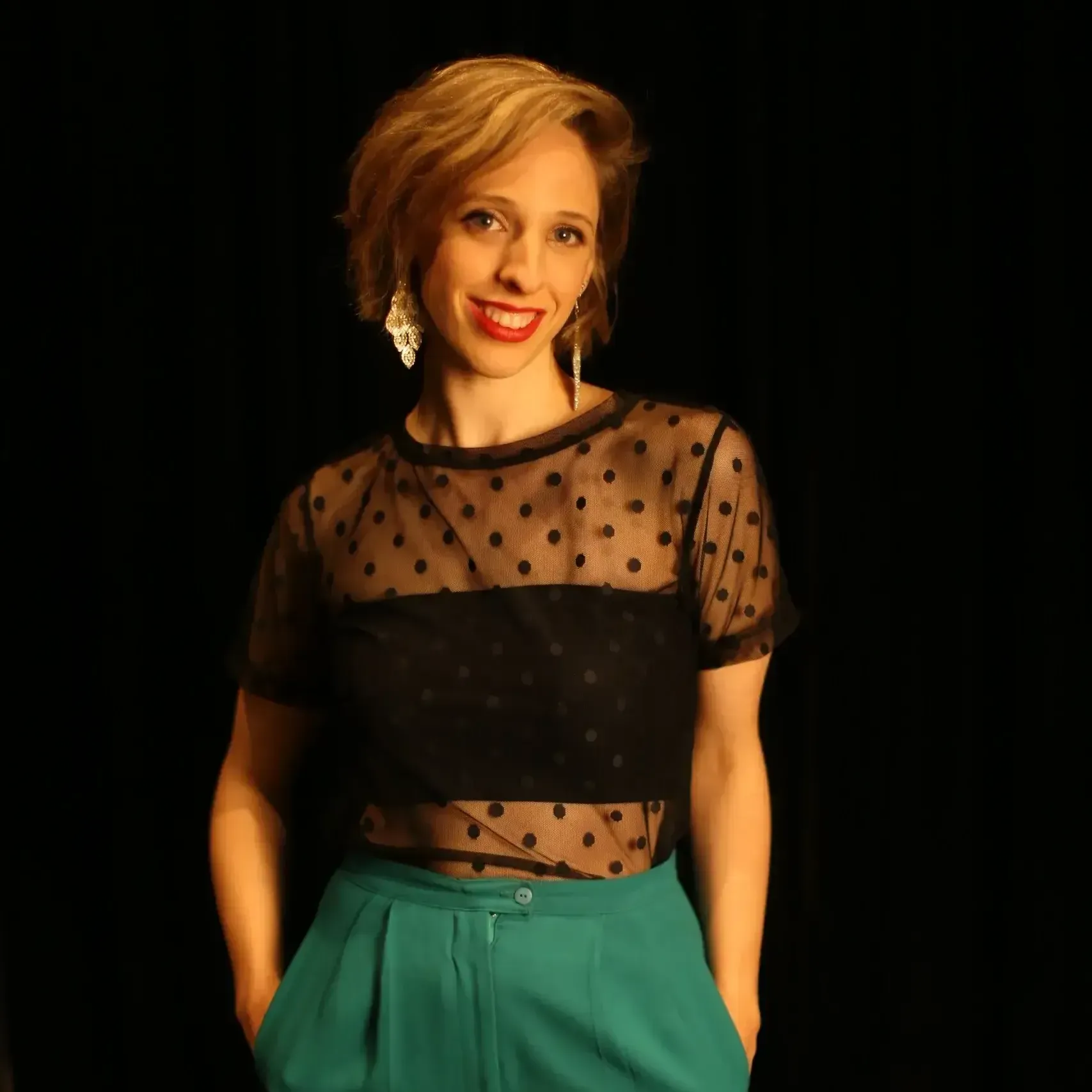 A person with short wavy hair, wearing a black polka-dot top and green trousers, posing against a black background.