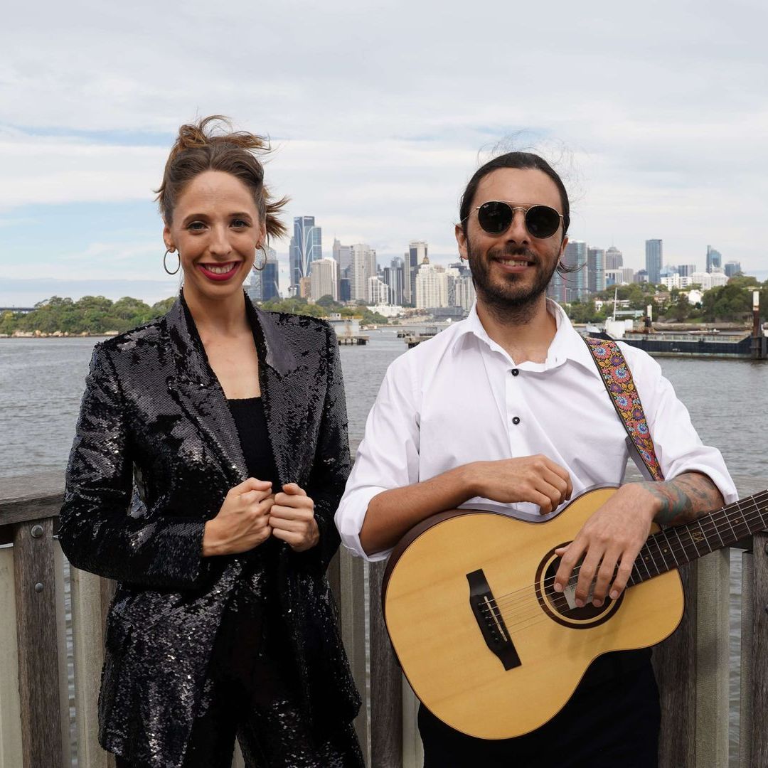A smiling duo poses by a city skyline and water; one person wears a sequined blazer, the other holds an acoustic guitar.