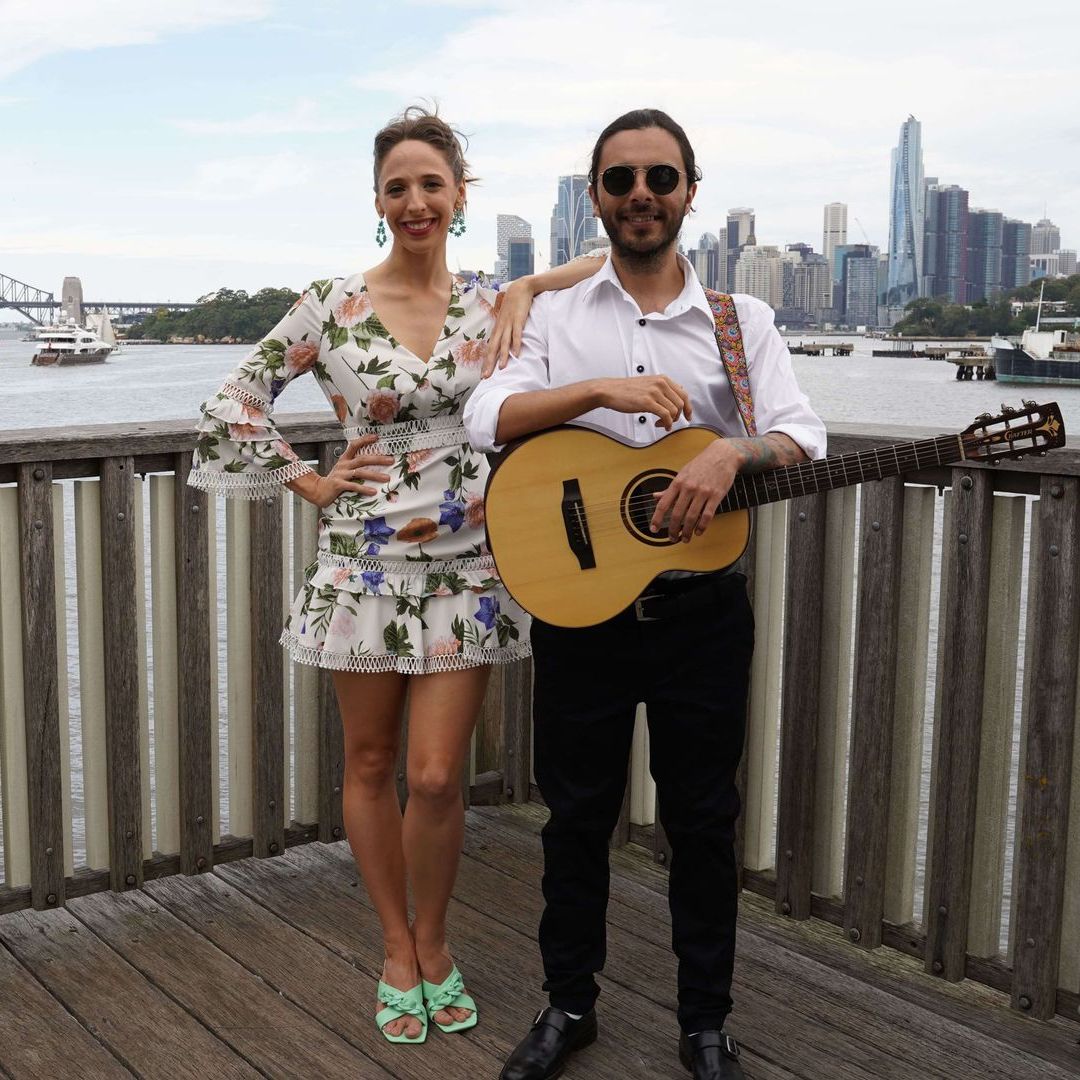A musician with a guitar and a woman in a floral dress pose on a boardwalk against a harbor and city skyline background.