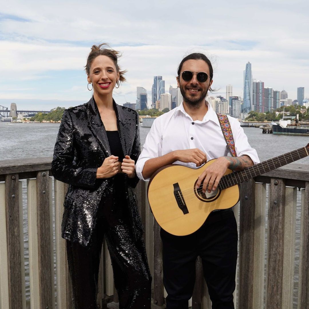 A musician with a guitar and a person in a sequin suit stand before a city skyline and water.