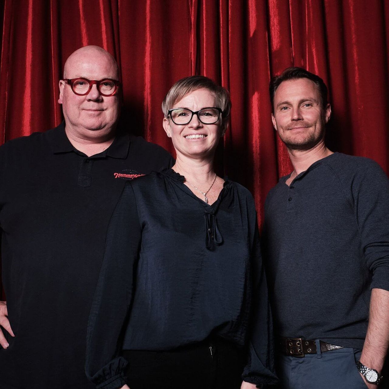 Three people standing in front of a red curtain, smiling and looking at the camera.