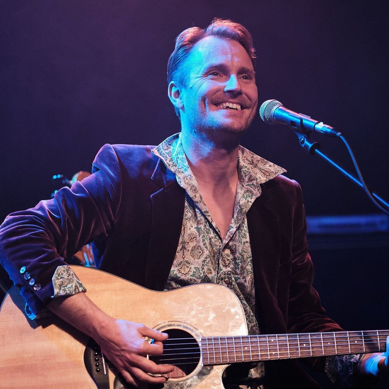 A smiling musician in a patterned shirt and dark blazer plays an acoustic guitar while performing into a microphone.