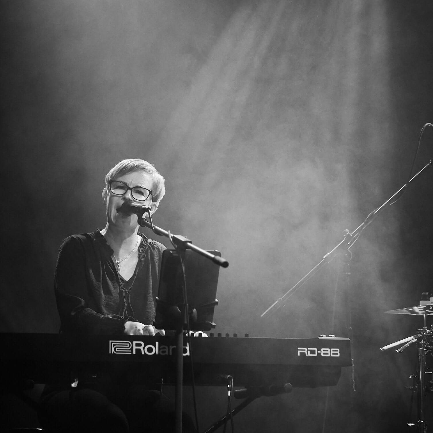 A person with short hair and glasses playing a Roland keyboard and singing into a microphone on a dimly lit stage.