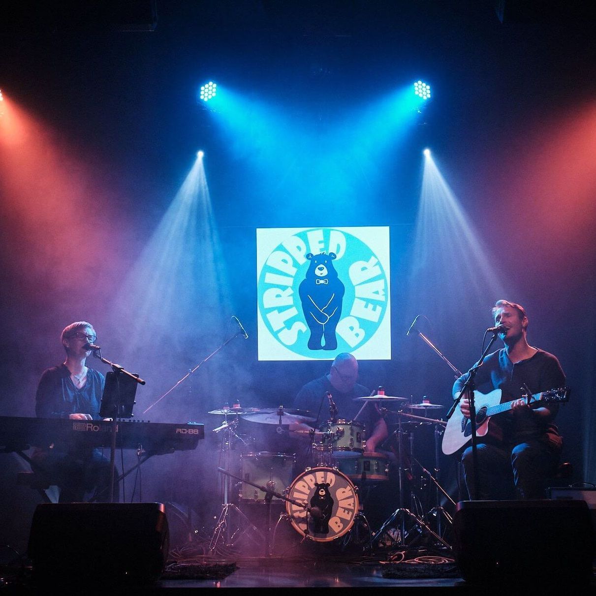 A three-piece band performs on a dimly lit stage under blue spotlights