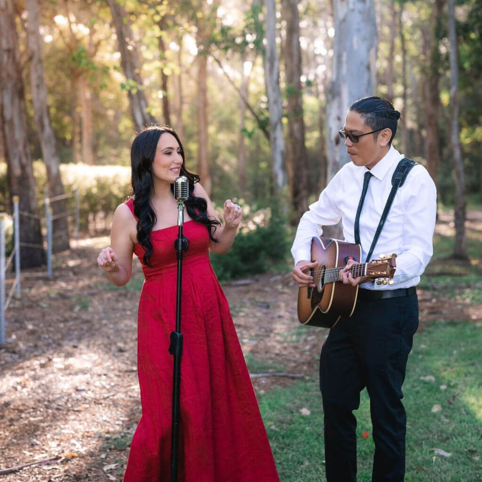 Two musicians perform outdoors in a sunlit wooded area, one singing and one playing acoustic guitar.