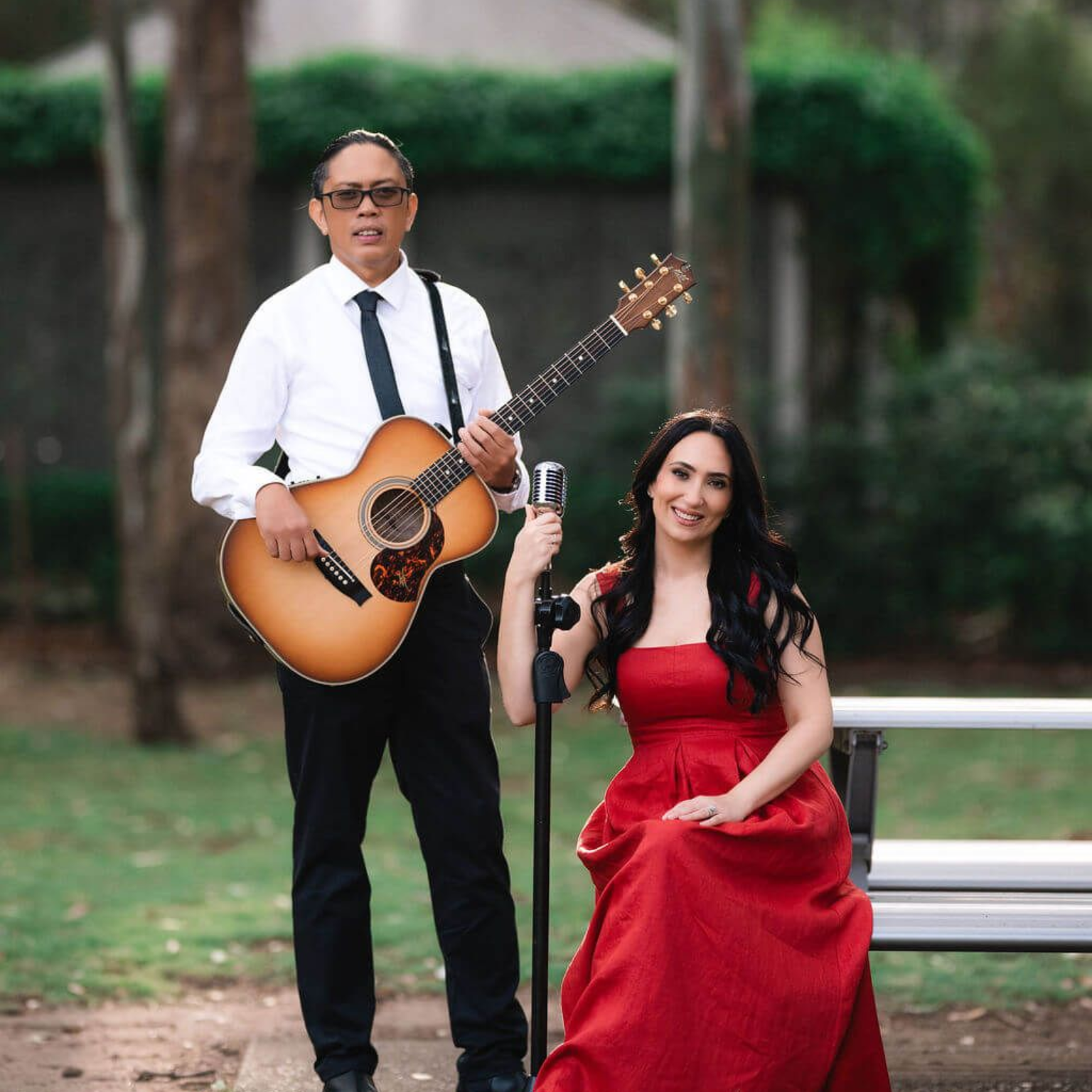 A man with a guitar stands beside a woman in a red dress seated by a microphone outdoors.