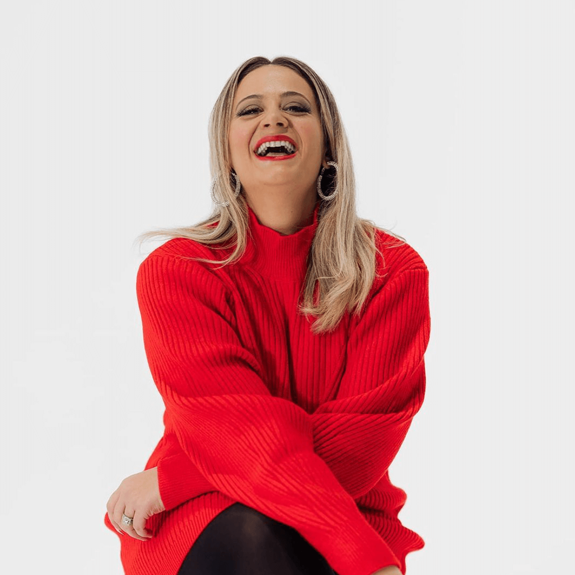 Woman in red sweater laughs, sitting against a white background.