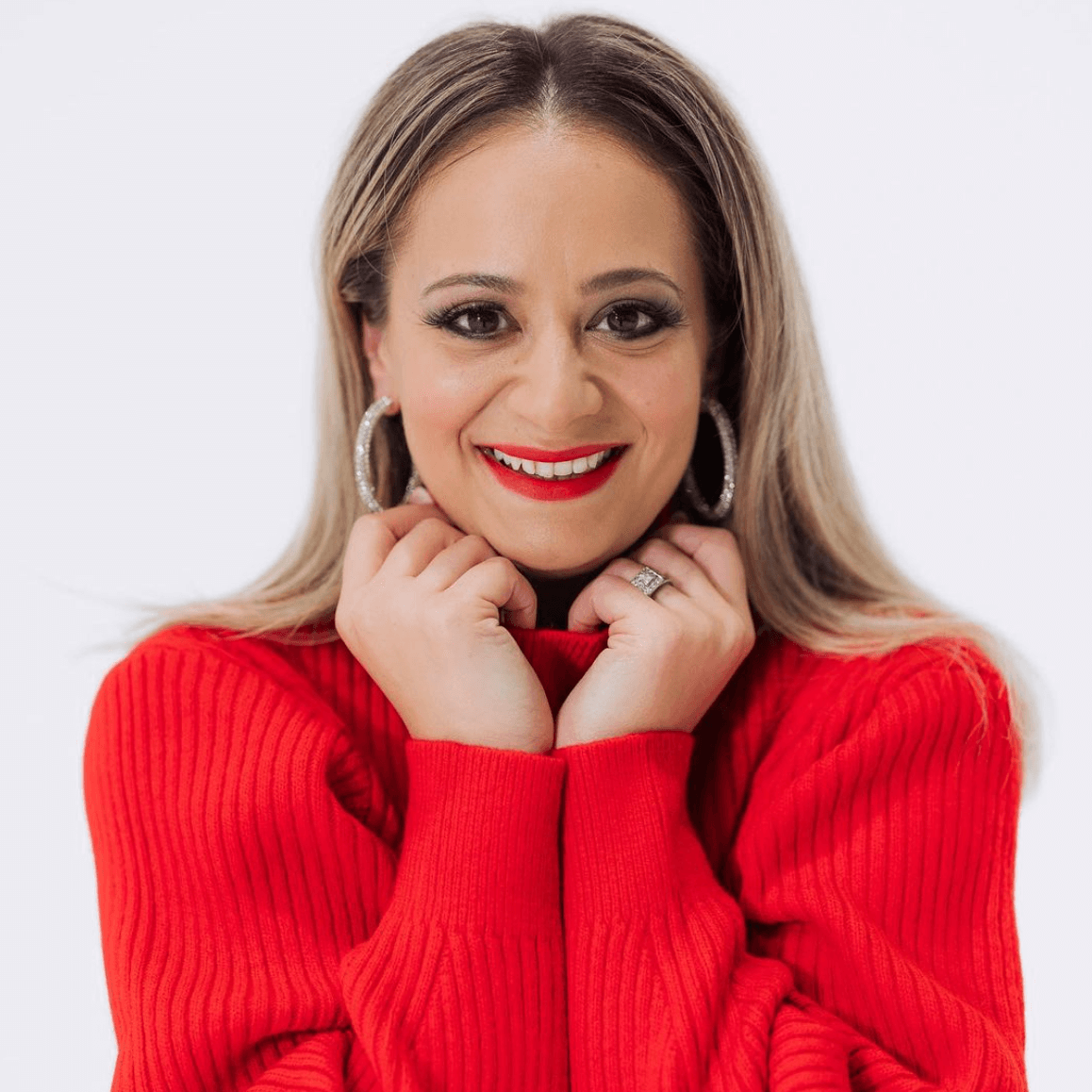 Woman with blonde hair, red sweater, smiling, hands near face, wearing large hoop earrings, red lipstick, white background.