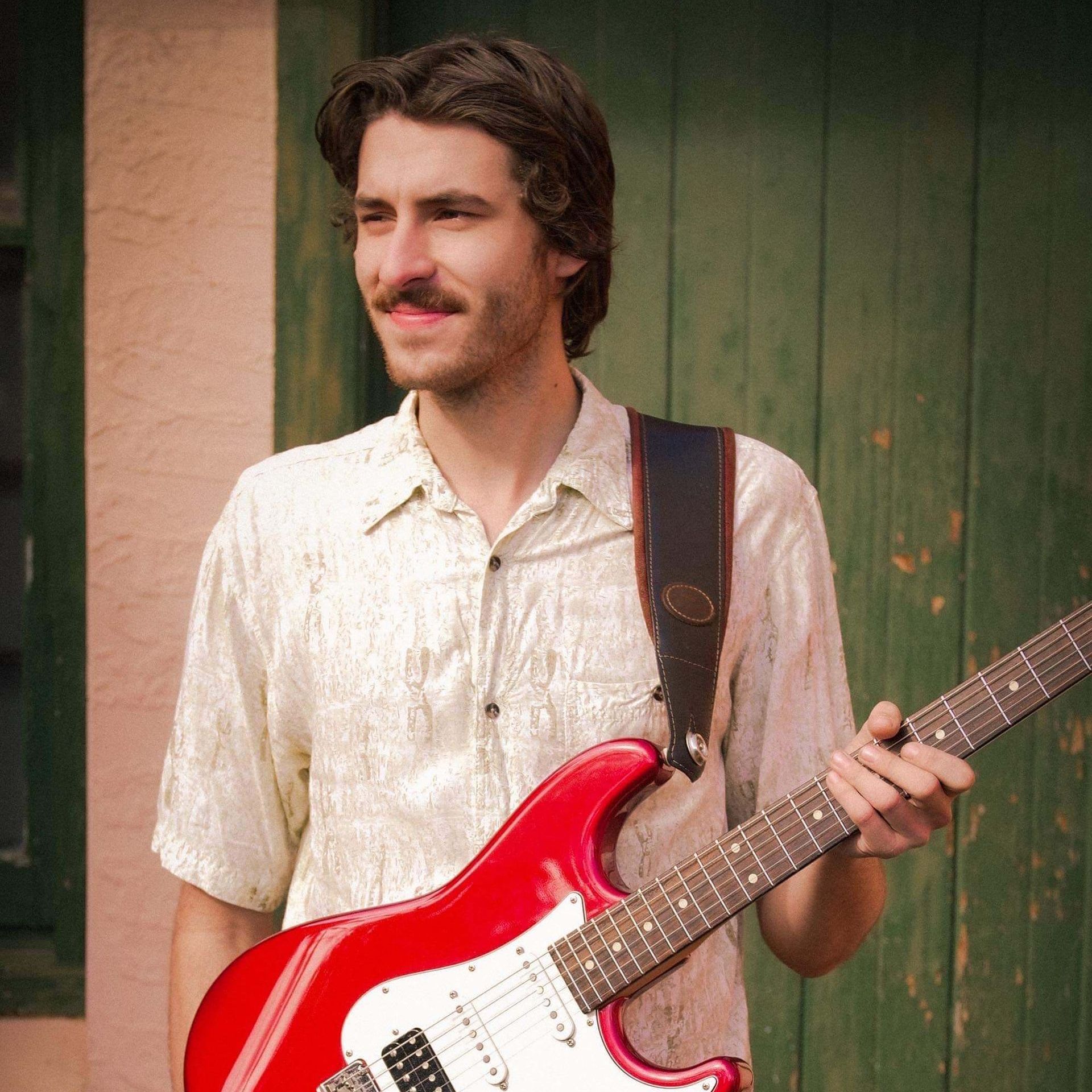 Man with a mustache, holding a red electric guitar, smiling, leaning against a green door.