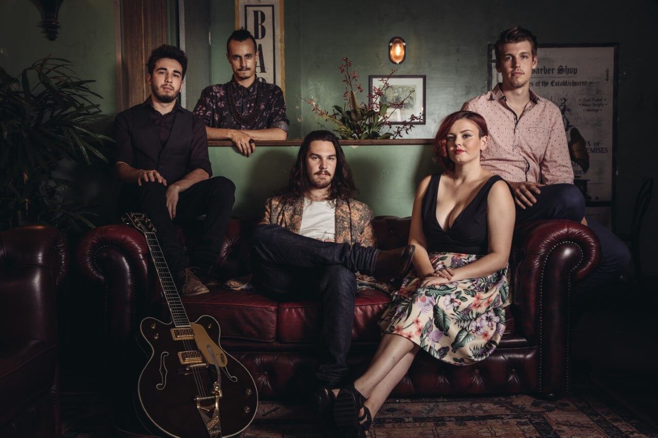 A band poses on and around a red leather couch in a dimly lit, vintage-style room with a guitar in the foreground.