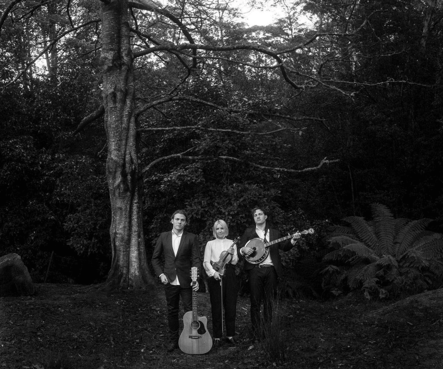 Three musicians standing in a forest, holding instruments. Black and white photo.