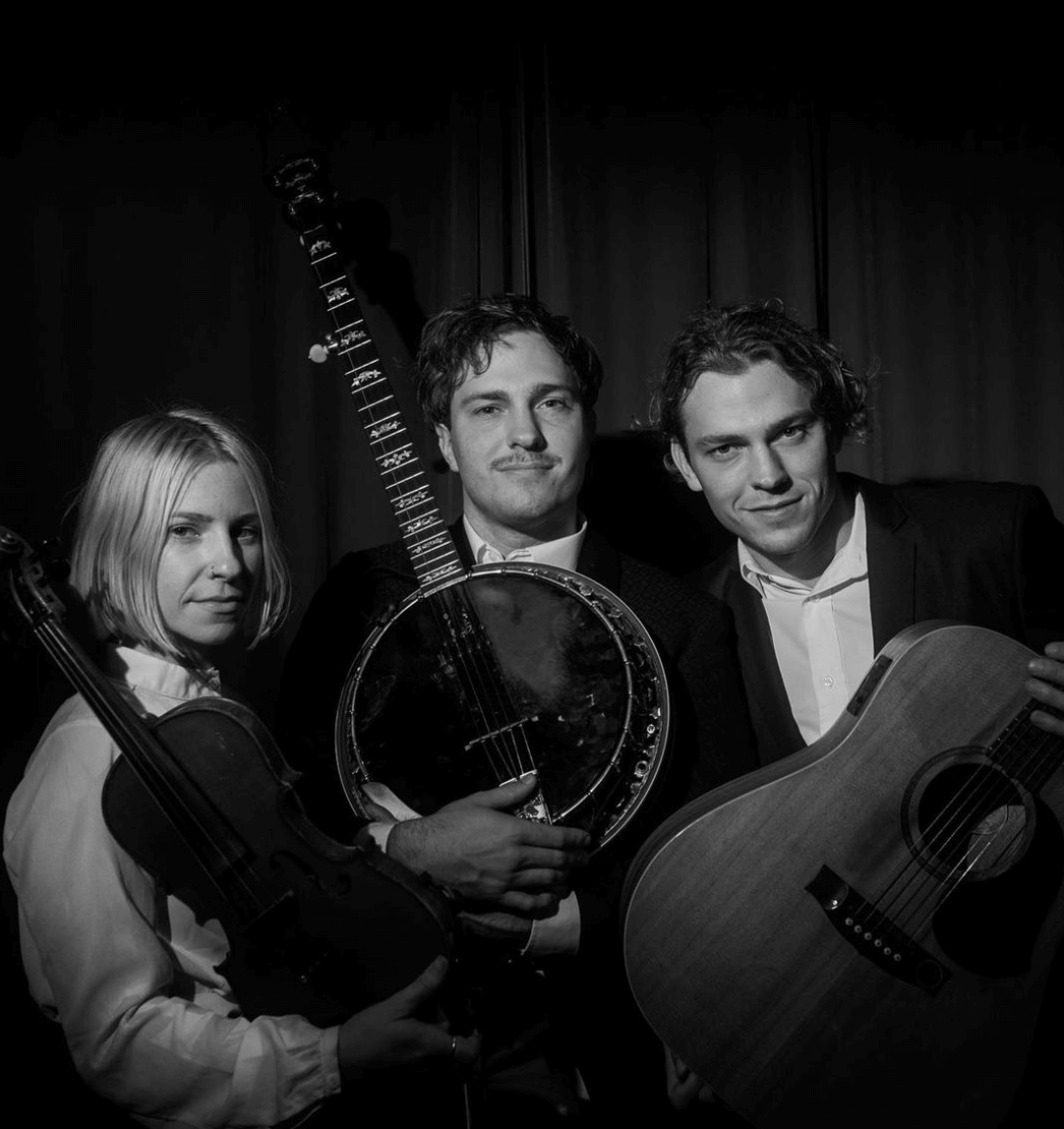 Three people with stringed instruments, smiling, in a dim setting.