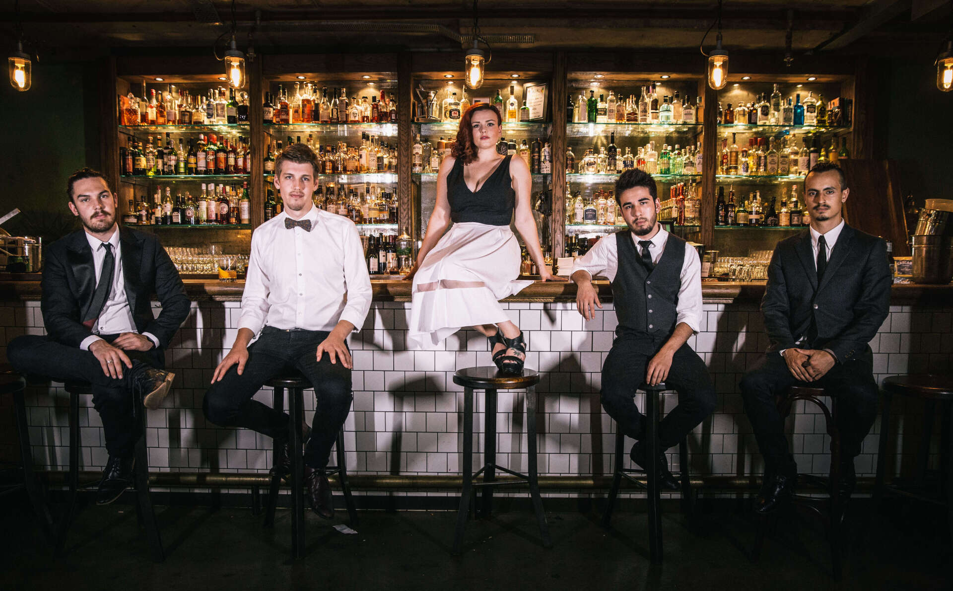 Four people in formal attire sit on bar stools in front of a well-stocked bar, posing for a group portrait.