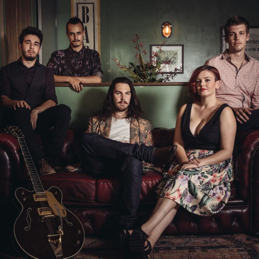 A band posing on a red leather sofa in a dimly lit bar, with one member holding a hollow-body electric guitar.