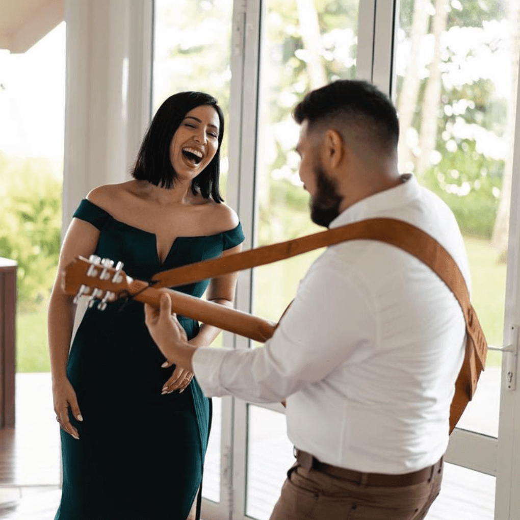 A person plays an acoustic guitar while a person in a dark green dress laughs near a large window.