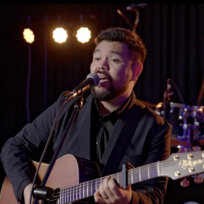 A musician wearing a black suit jacket sings into a microphone while playing an acoustic guitar on a dark stage.