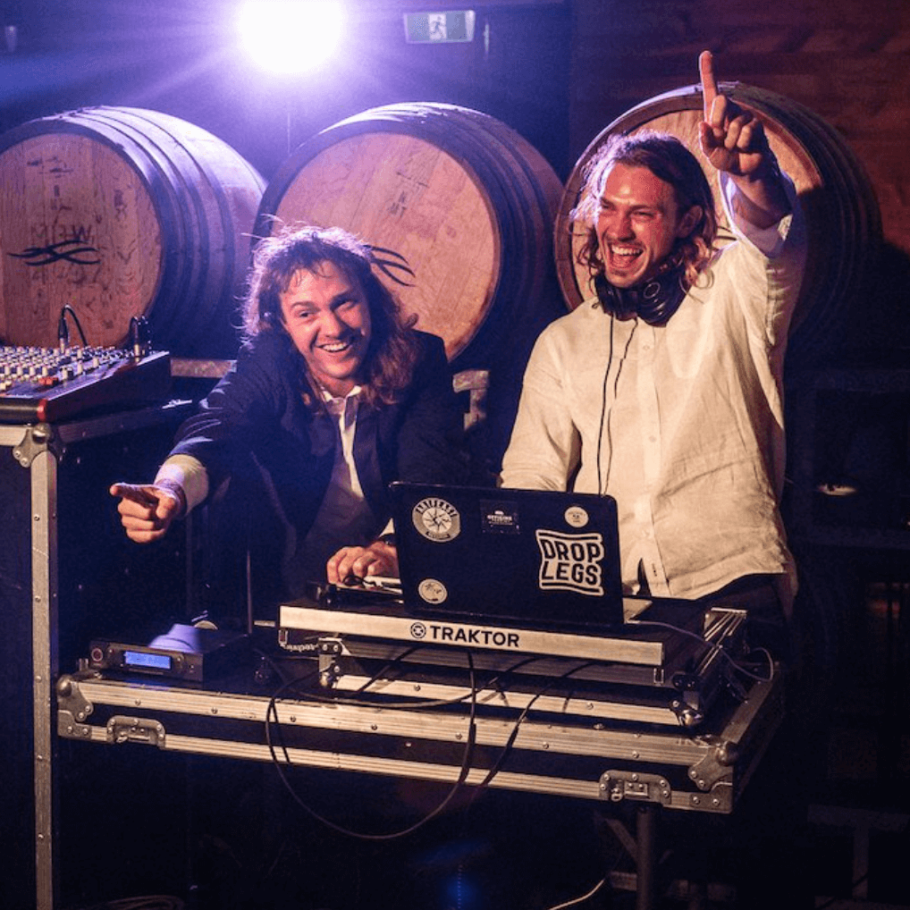 Two DJs with long hair smiling and gesturing, behind a DJ setup, with barrels in the background.