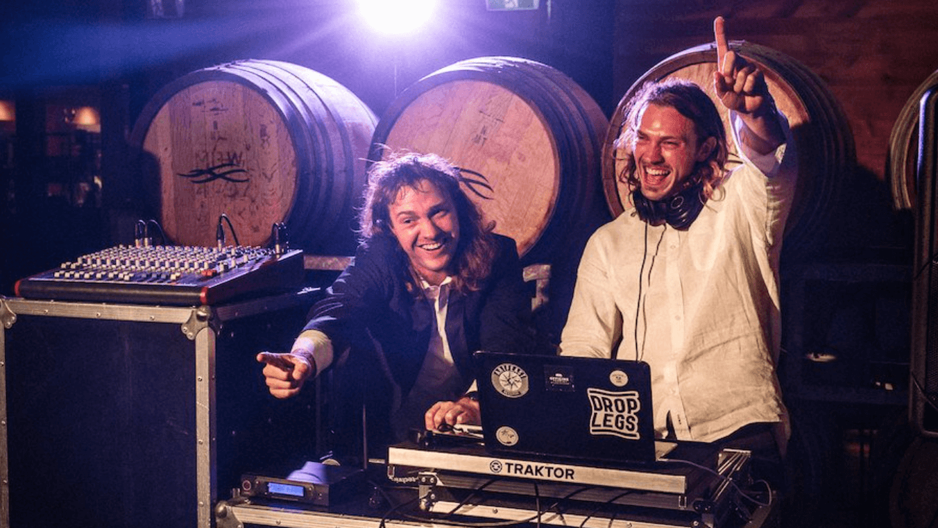 Two men are playing music on a laptop in front of barrels.