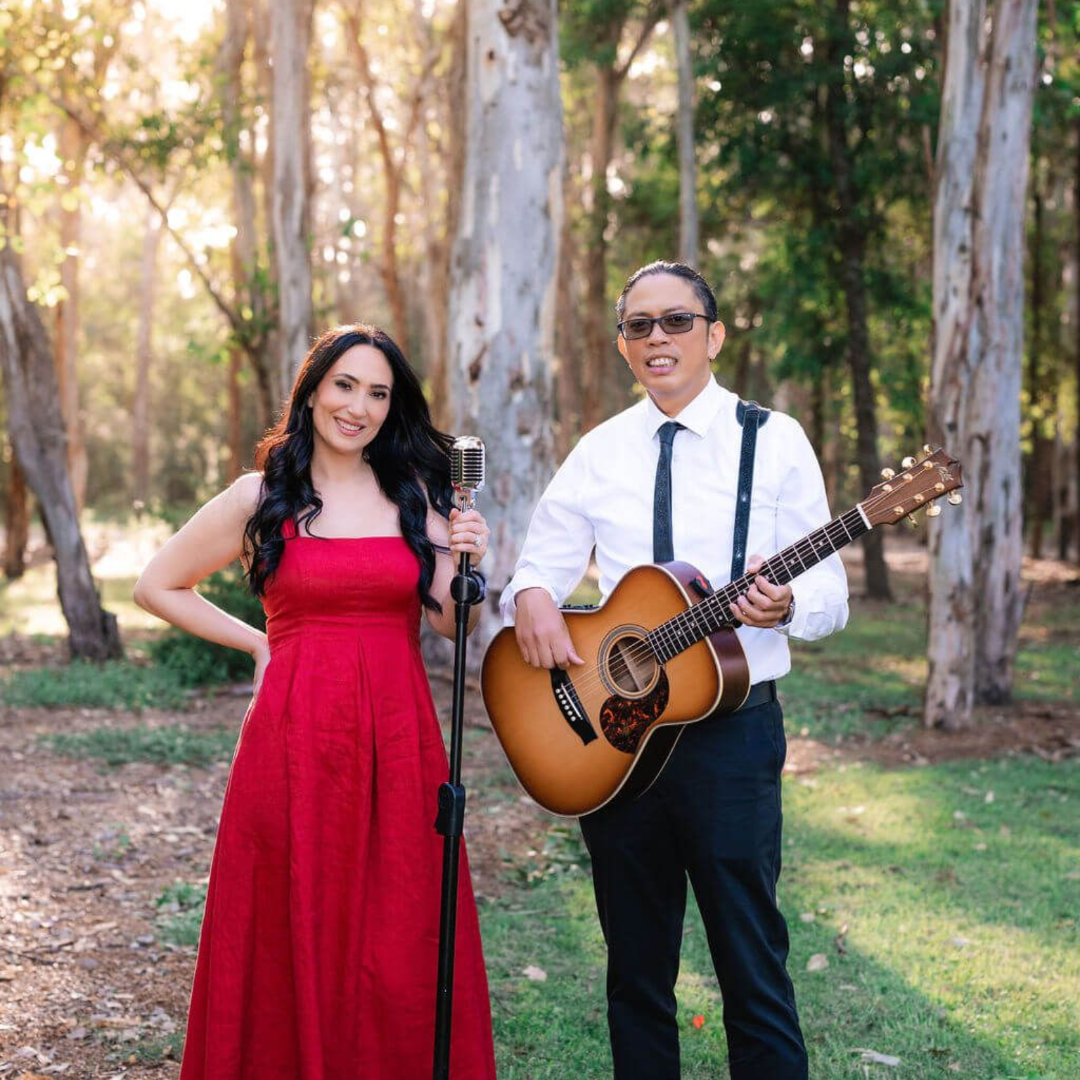 A duo performs in a forest, with a person in a red dress by a microphone and a musician playing an acoustic guitar.