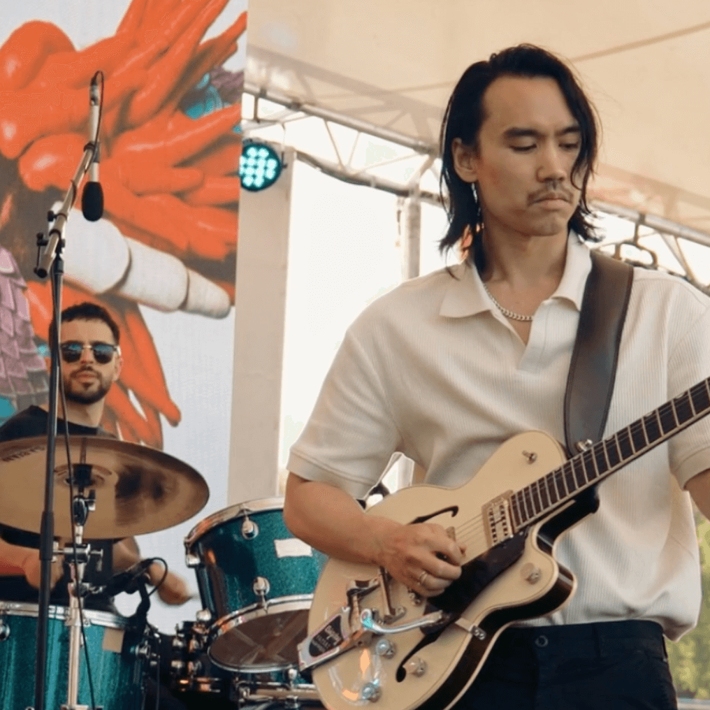 Band playing live outdoors. Guitarist in white shirt, drummer behind him. Stage setup, colorful backdrop.