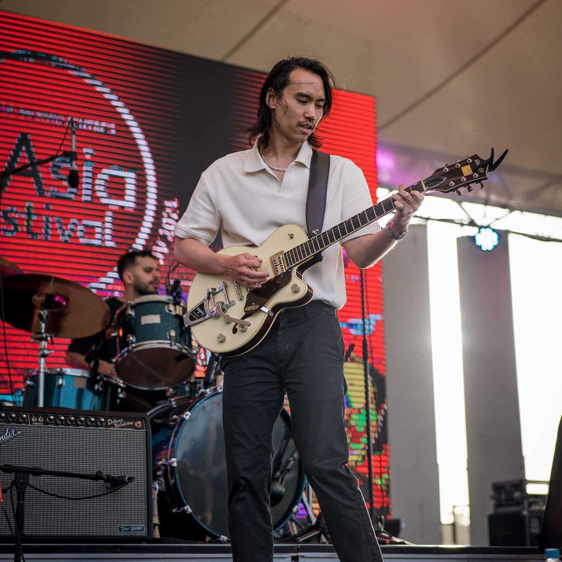 Man playing electric guitar onstage at a festival, with a drummer visible behind him. Red backdrop.