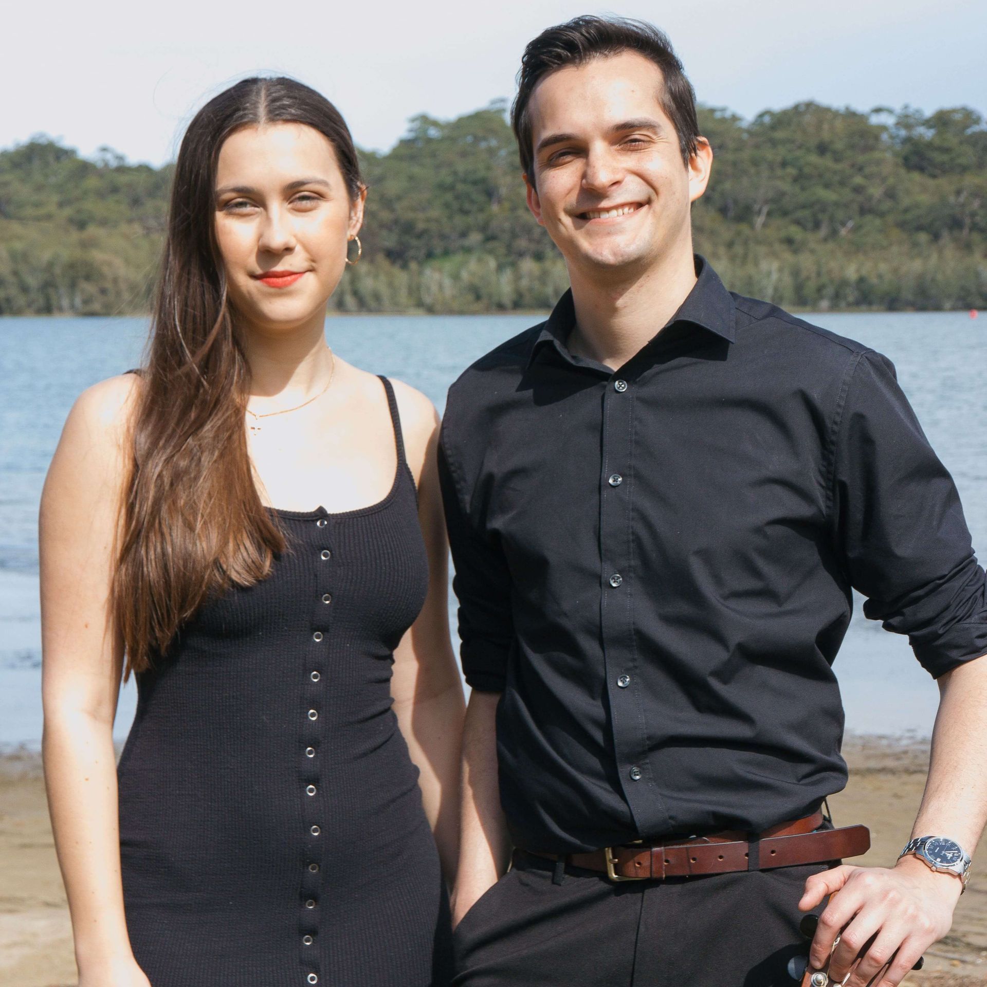 A couple in black clothing stands side-by-side, smiling, with a lake and forest in the background.