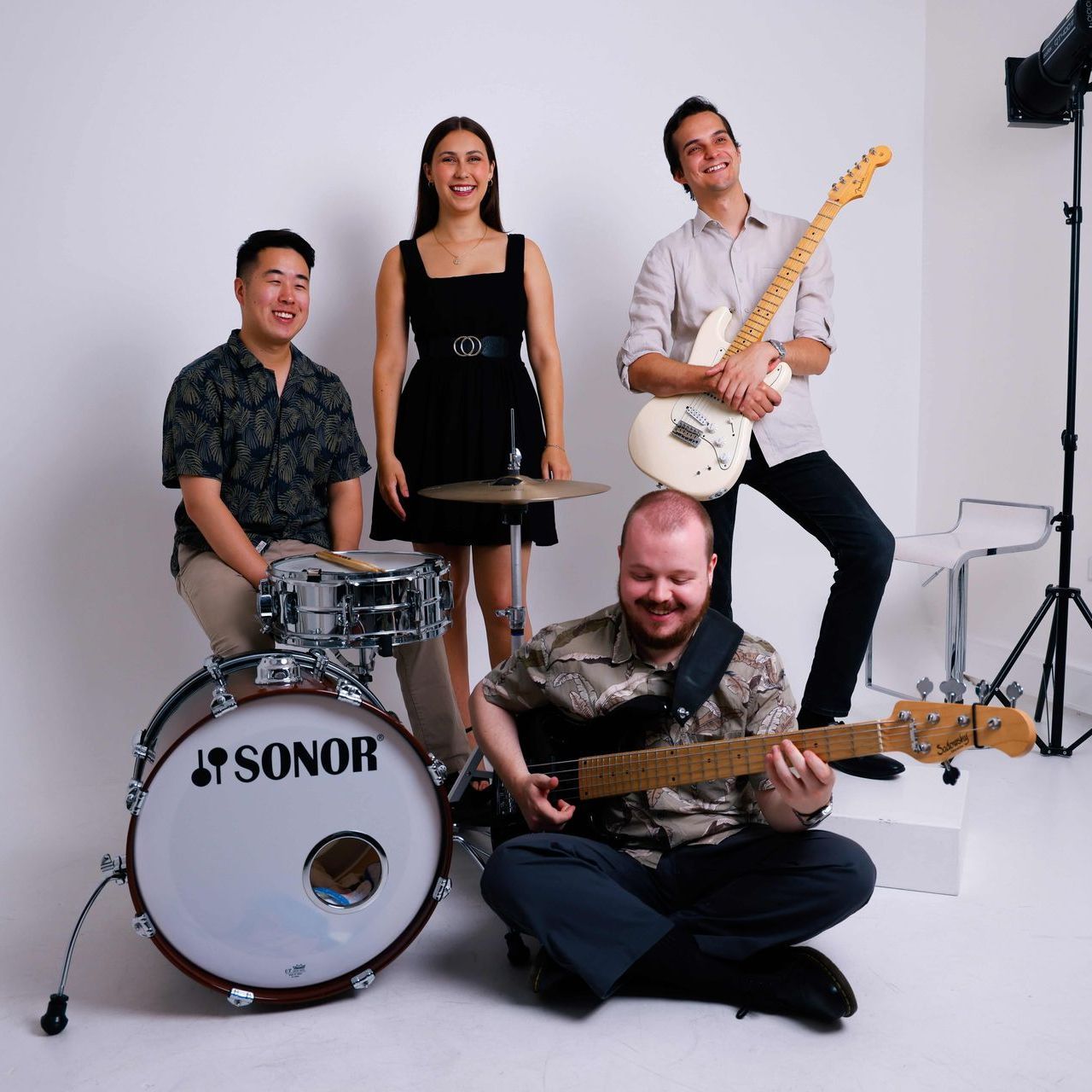 Four musicians posed in a studio with drums and guitars. Three stand behind a seated member holding a bass guitar.