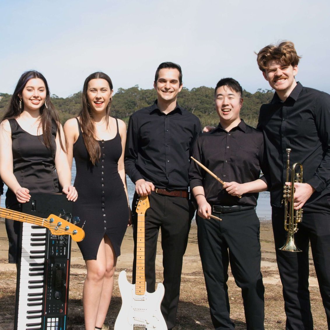 A group of five musicians stands outdoors in black clothing, holding musical instruments including a keyboard, guitar, and trumpet.