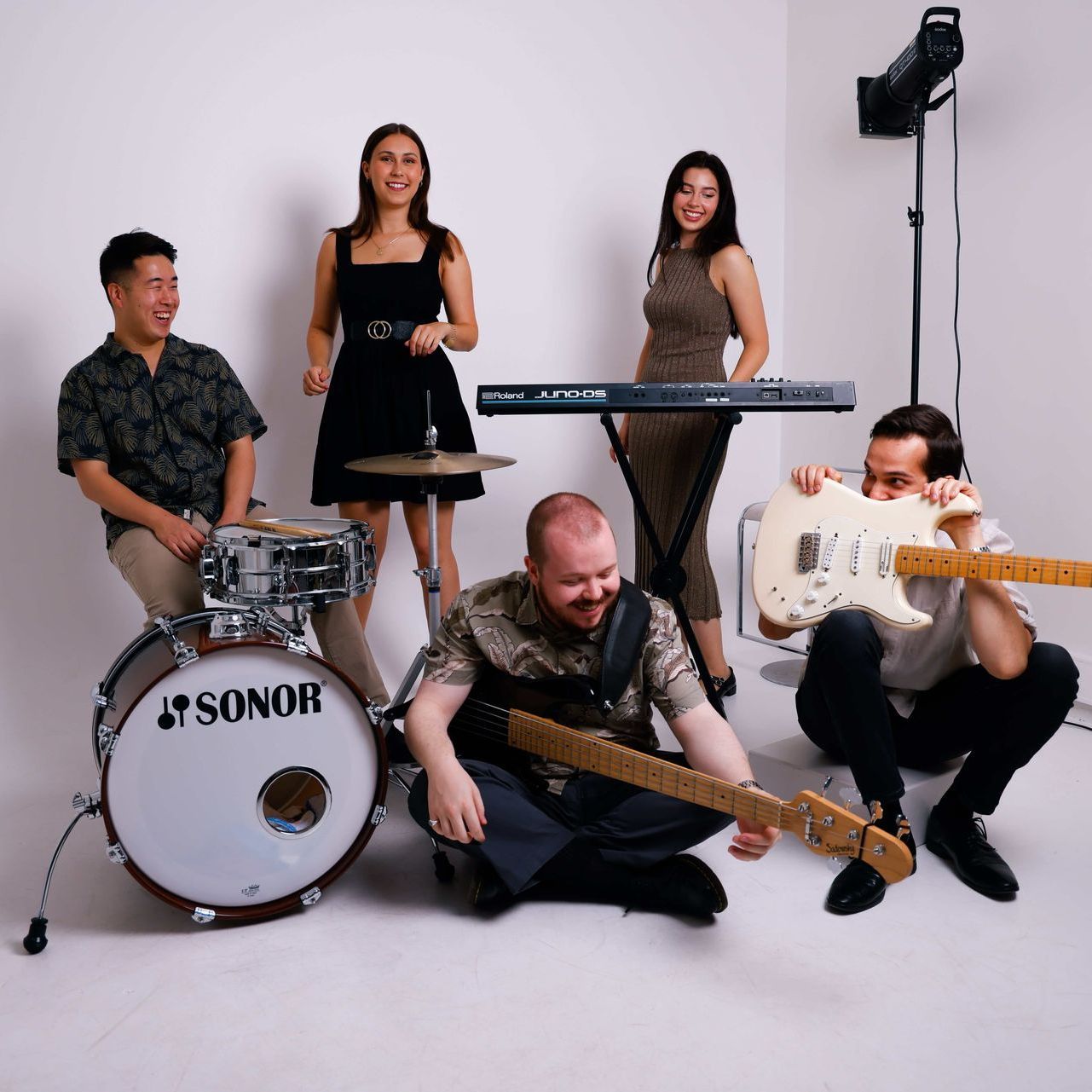 Five band members posing in a studio with drums, guitars, and a keyboard, all laughing and smiling.