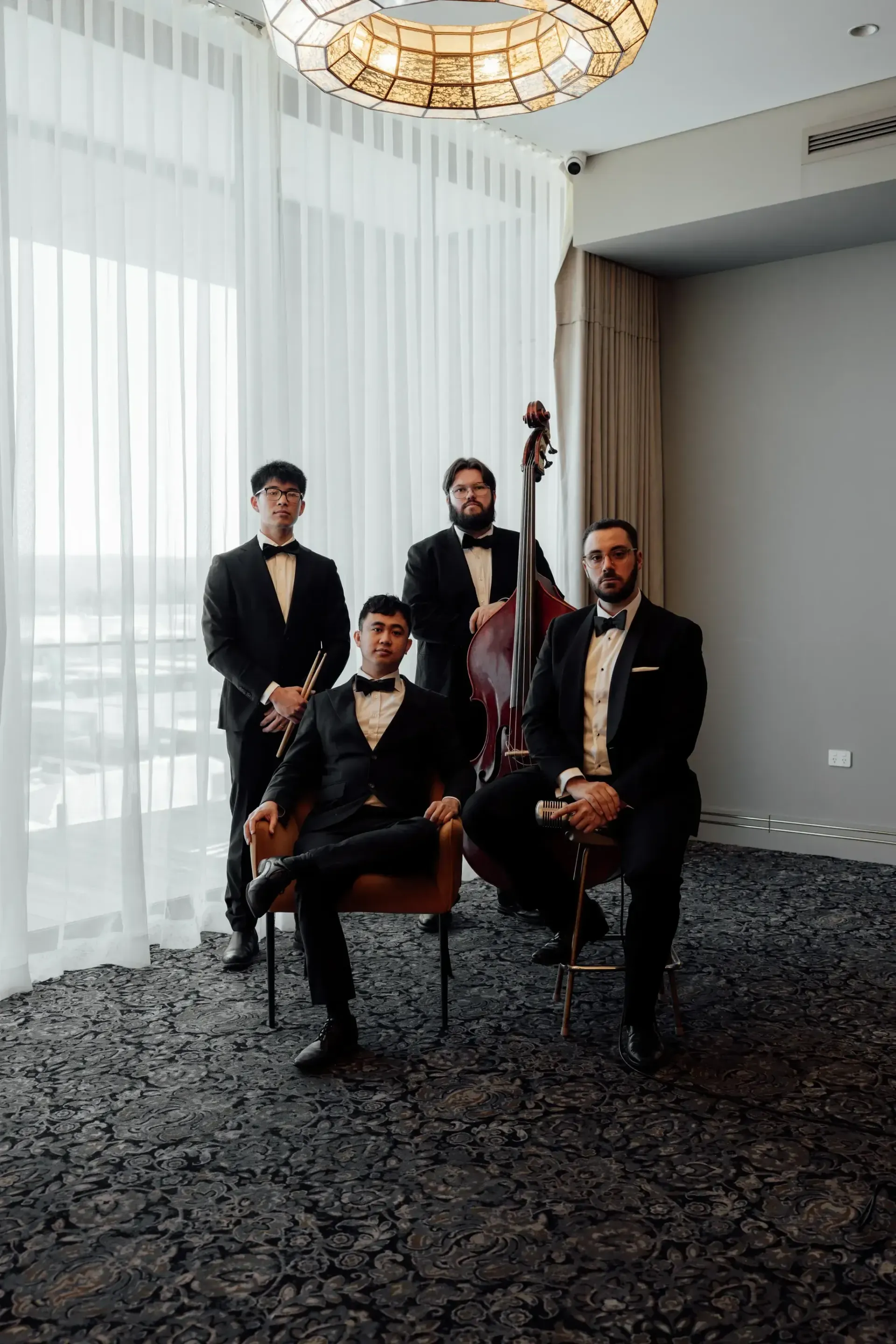 Four musicians in black suits posing with string instruments in a bright room with tall curtains.
