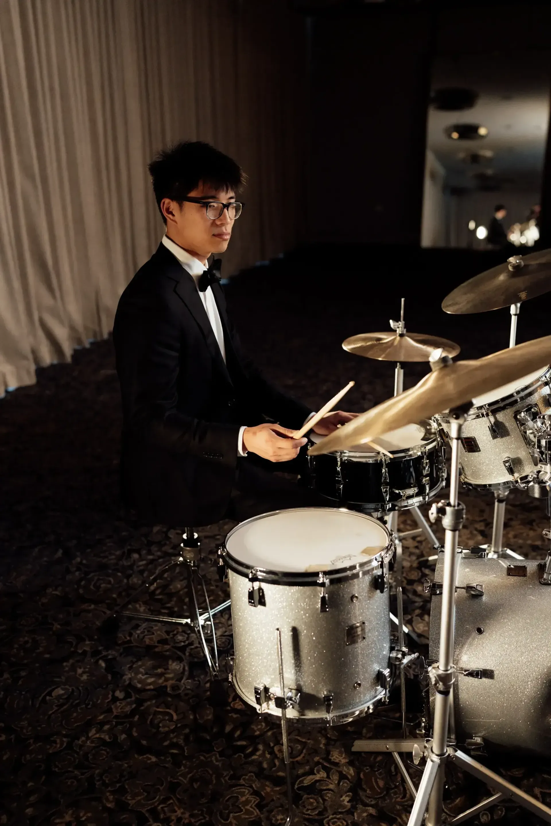 Drummer in a dark suit playing a drum kit on a dimly lit stage