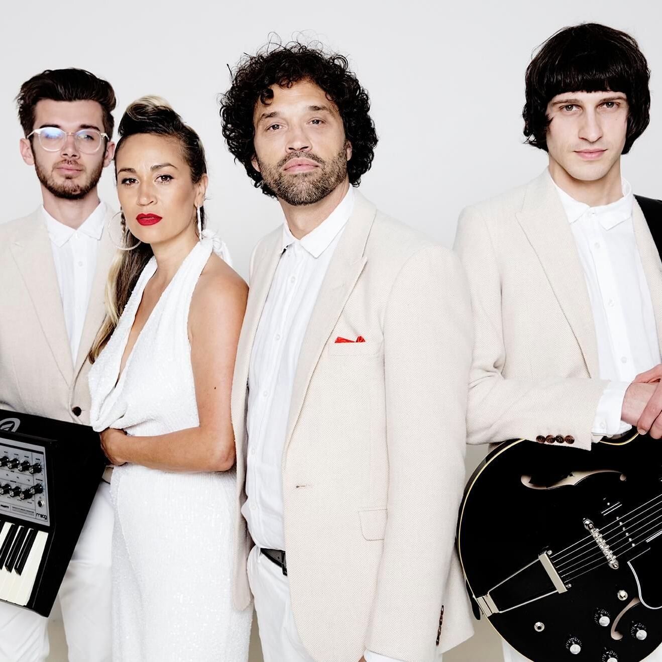 Four people in cream-colored suits pose for a photo. A woman in a white dress stands beside a keyboard.