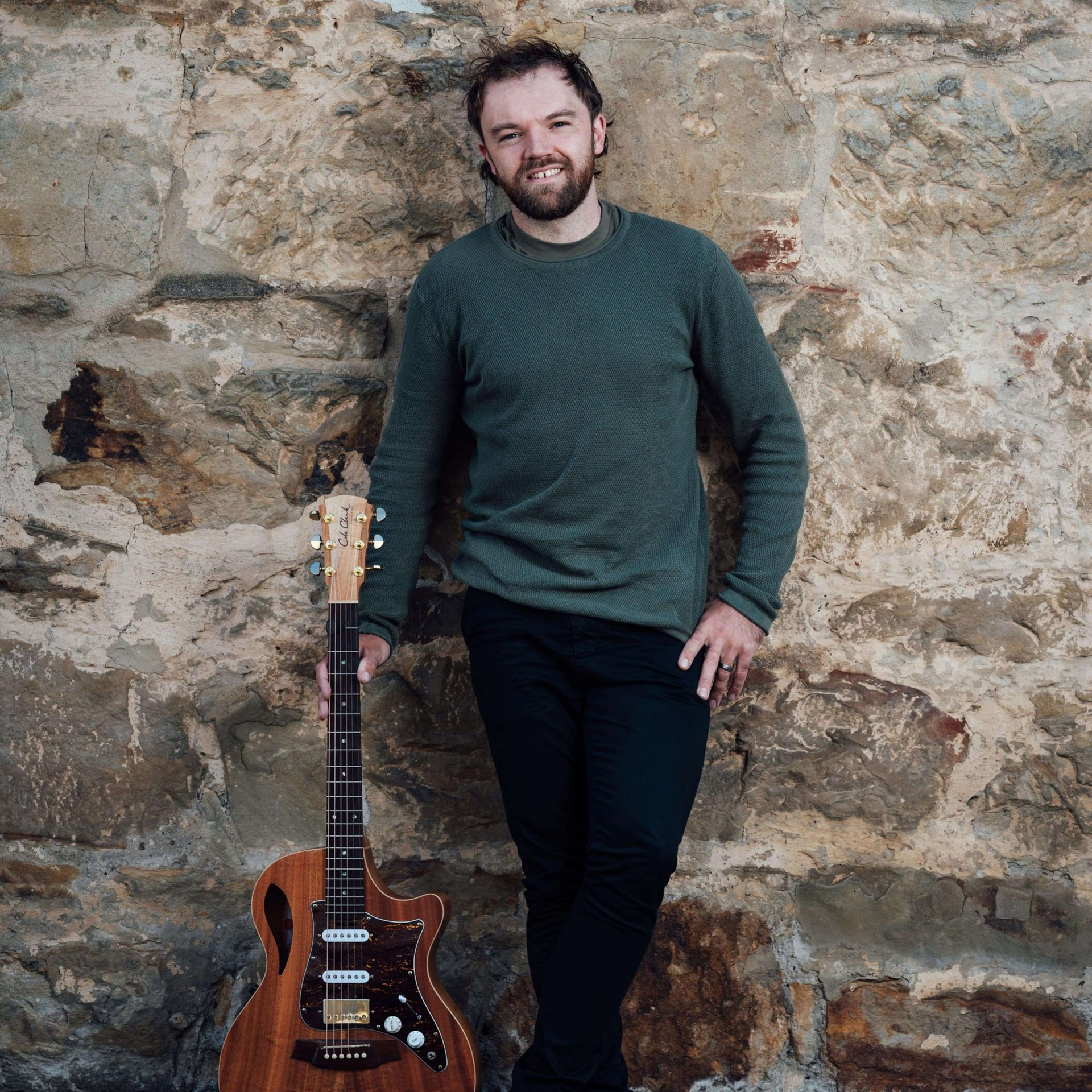 Man leans against a stone wall, holding a brown guitar. He wears a green sweater and black pants.
