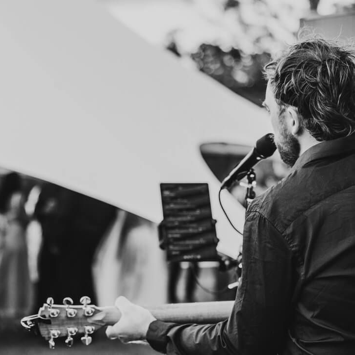 Man playing guitar and singing into a microphone, outdoors. Black and white photograph.