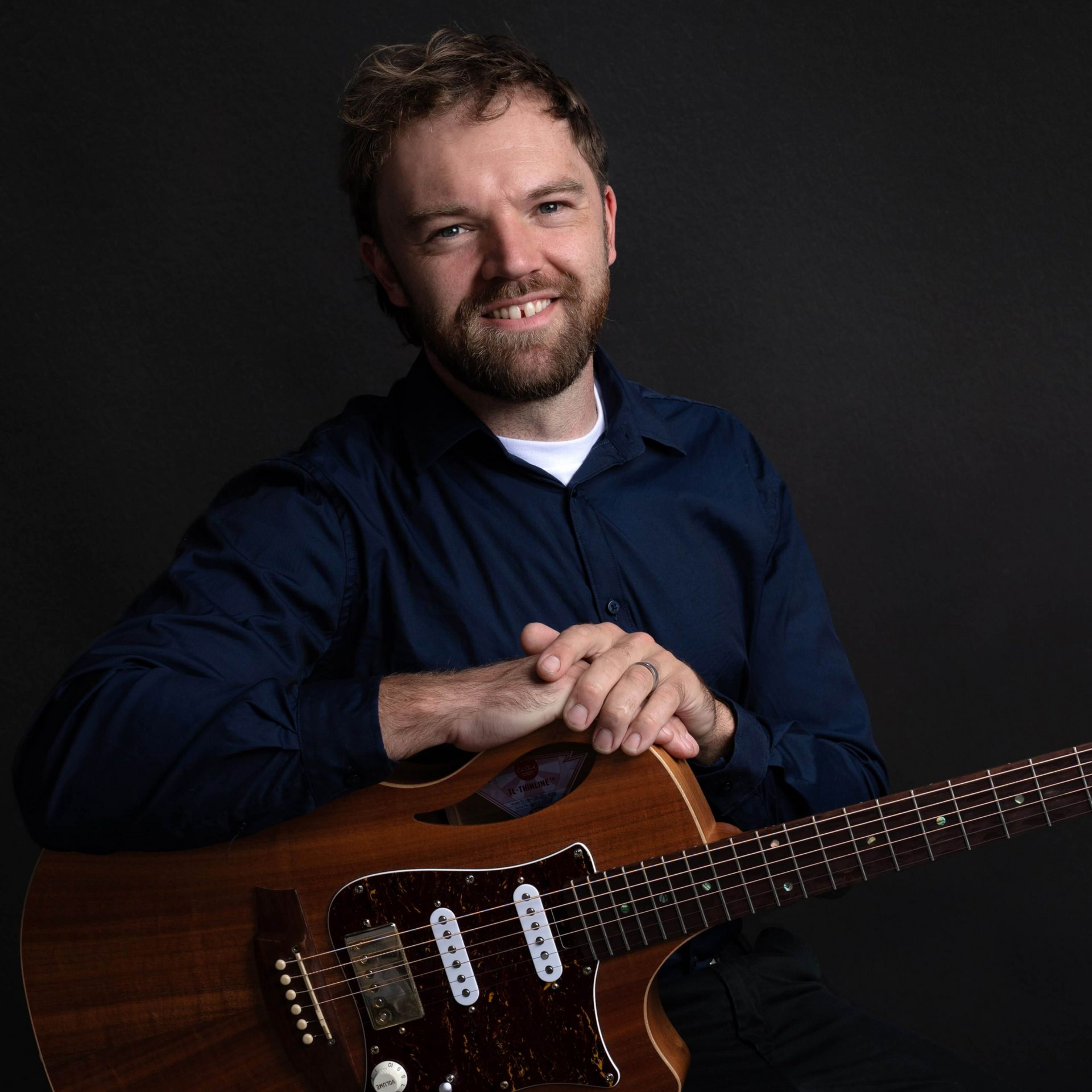 Man holding a brown electric guitar, smiling, dark blue shirt, dark background.