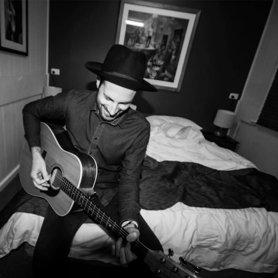 Man in a fedora plays acoustic guitar, smiling, sitting on a bed in a room. Black and white photo.