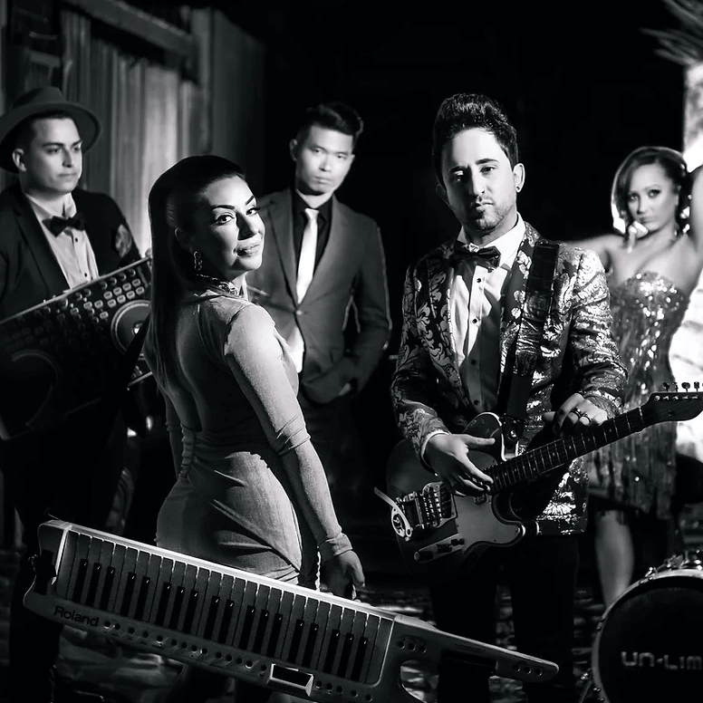 Band posing in black and white. Singer in dress, guitarist, keyboardist, drummer, setting is a wooden backdrop.