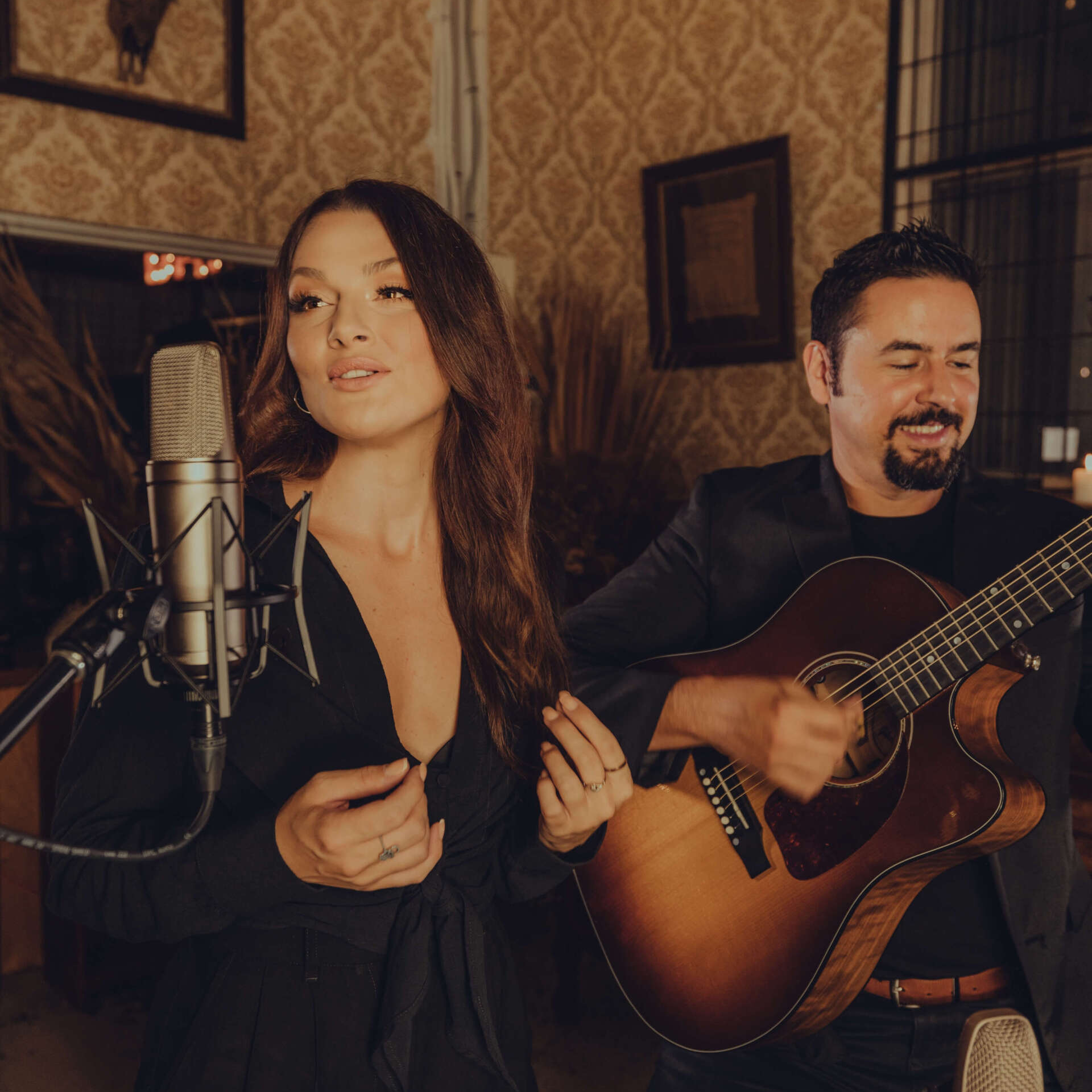 Woman singing into microphone; man playing acoustic guitar in a room with a vintage aesthetic.
