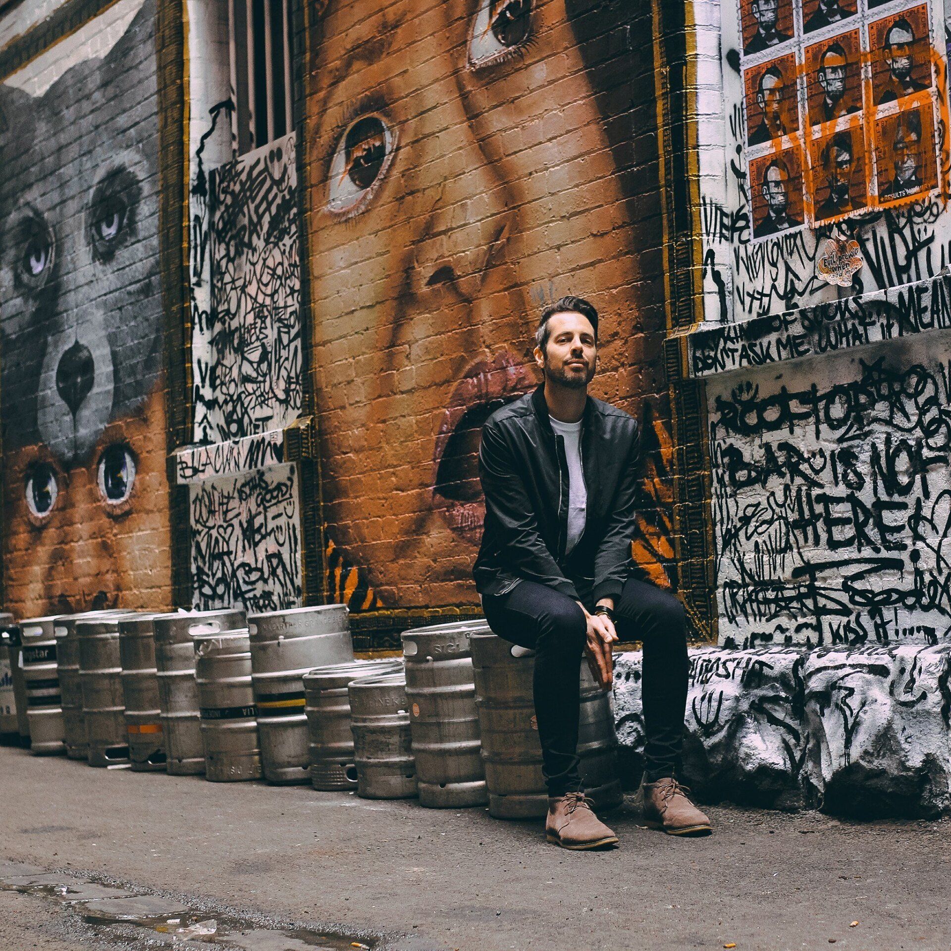 Man sitting on beer kegs, looking upward. Mural background with graffiti and dog and face paintings.